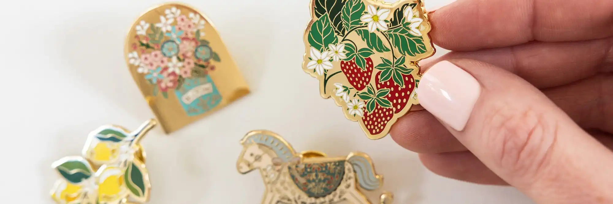 Assortment of enamel pins, including strawberry, lemon, floral, and rocking horse designs.