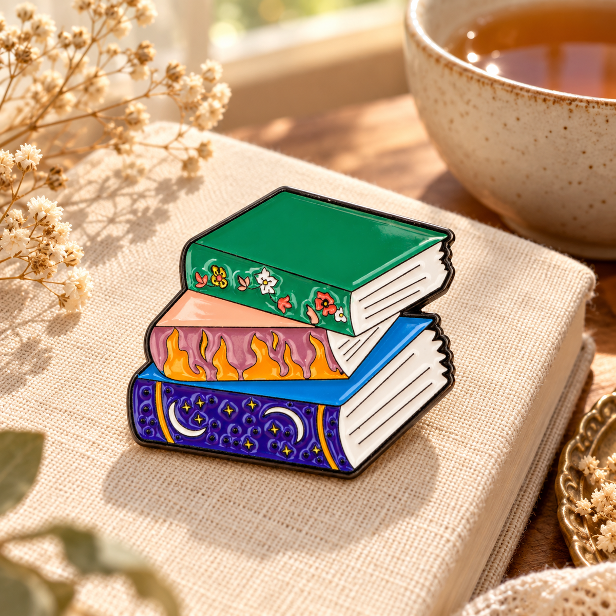 Books enamel pin on a cozy linen book beside tea and flowers