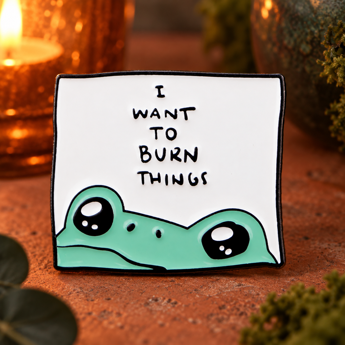 I Want to Burn Things frog enamel pin on a warm terracotta table