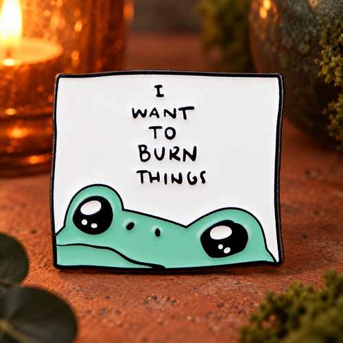 I Want to Burn Things frog enamel pin on a warm terracotta table