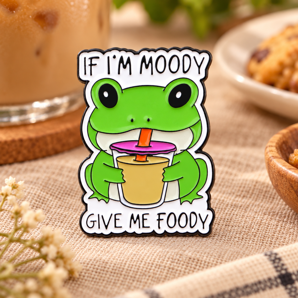 If I'm Moody Give Me Foody frog enamel pin in a cozy café setting