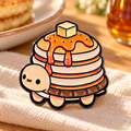 Pancake Stack Turtle enamel pin on a sunlit breakfast table