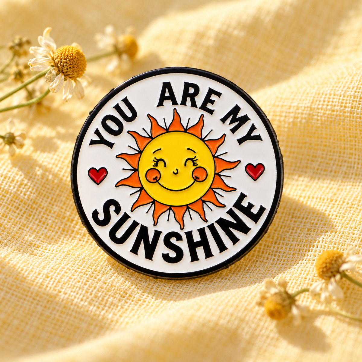You Are My Sunshine enamel pin displayed on warm yellow linen with chamomile flowers