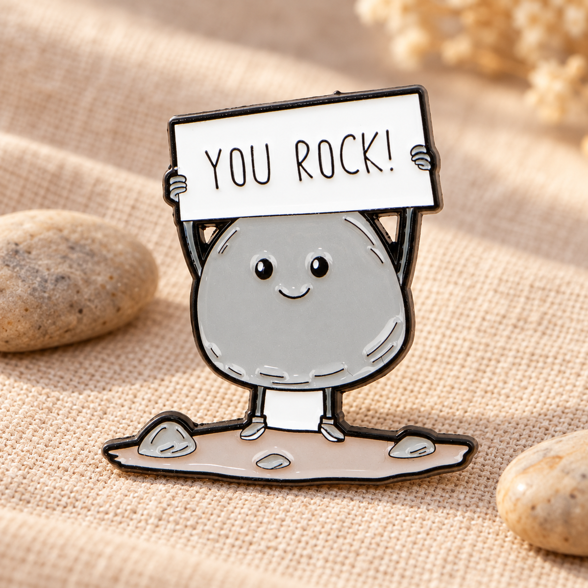 You Rock enamel pin displayed on warm linen with smooth pebbles